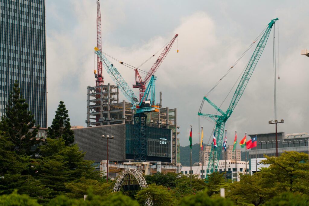 Cranes tower over a construction site amidst Taipei's urban landscape, featuring flags and a modern skyline.
